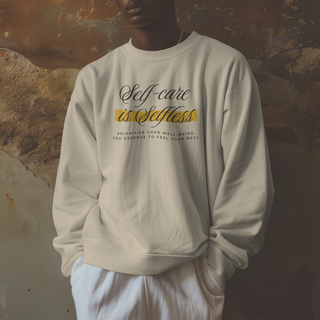 A person wearing a cream-colored crewneck sweatshirt with a graphic print that reads 'Self-care is Selfless' in black and yellow text, paired with white sweatpants, set against a rustic, textured wall.